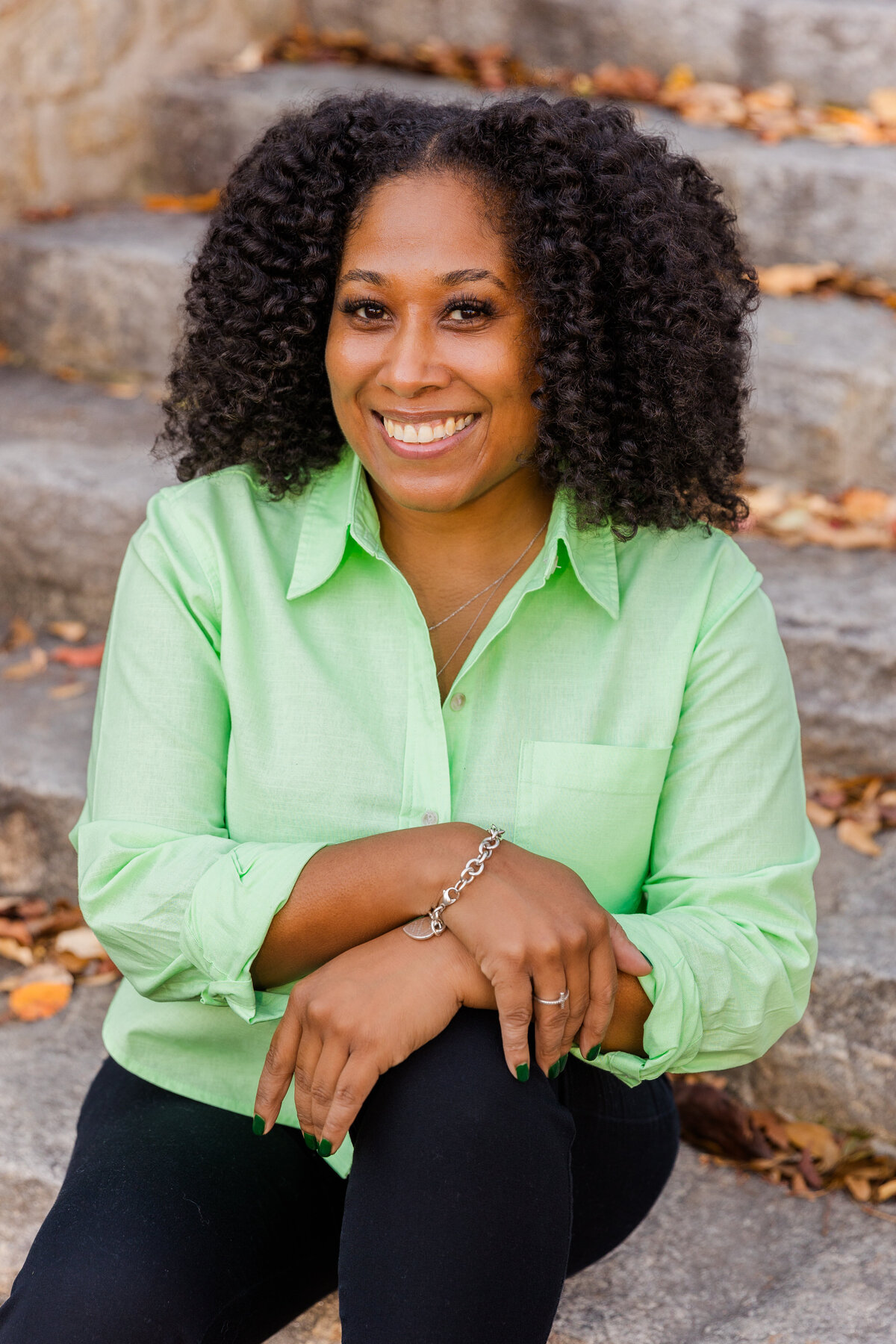 woman wearing a black pant anf a light green blouse sitting on outdoor stairs wrists croosed above the knee during portraits session