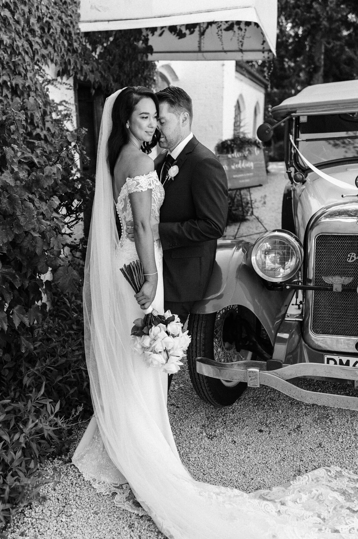 black and white photo of bride in off the shoiulder lace dress with groom in front of vintage car at trent vineyard at prebbleton