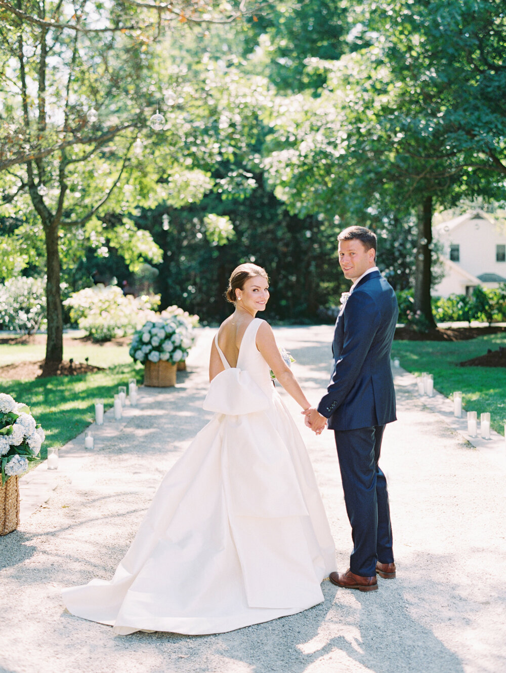 The delightful scene captures a heartwarming moment at a waterfront estate tented wedding in The Hamptons, New York, immortalized by the skilled lens of photographer Bonnie Sen, complemented by East West Films as Videographers. Amidst the sun-dappled surroundings, a radiant bride and dashing groom stand hand in hand, their eyes gleaming as they gaze back at the camera. The bride's exquisite white gown, complete with a graceful train and a delicate bow in the back, harmonizes beautifully with the groom's debonair look in a sharp dark suit. The harmonious couple is embraced by lush greenery and vibrant floral displays, adding an extra touch of elegance to the enchanting setting.
