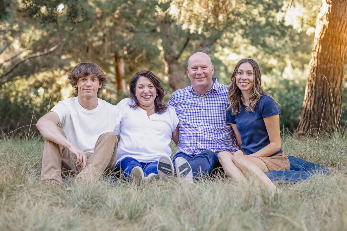 A family of four with brown hair wearing blue and white sitting on a navy picnic blanket in the grass with their arms around each other, smiling at the camera.