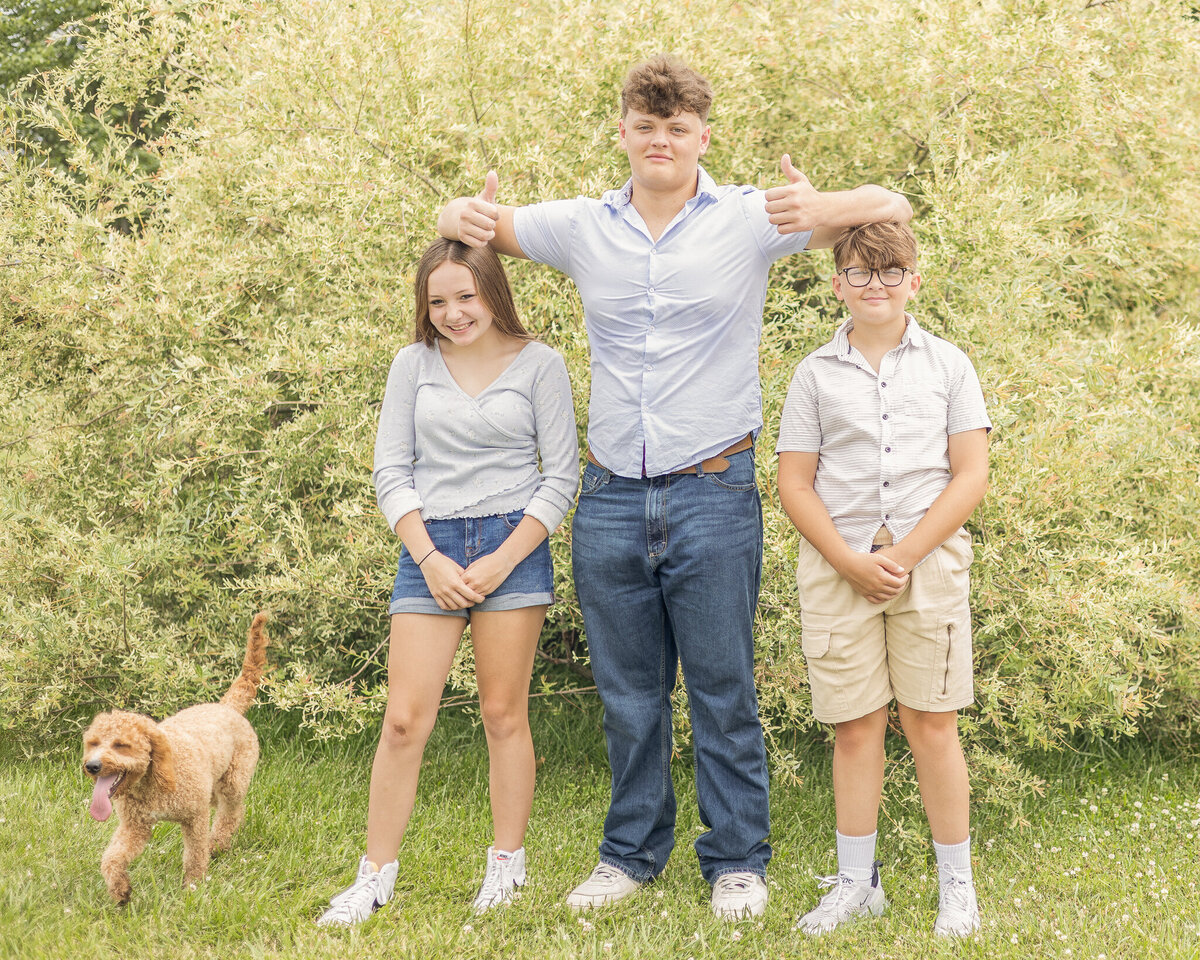 Two brothers and a sister standing in the grass in front of a bush.  Oldest is wearing a light blue button up shirt, blue jeans, white shoes, closed smiling at the camera, and has his arms on his siblings heads giving thumbs up.  Younger brother is wearing a light tan button up shirt, khaki shirt, white shoes, and smiling at the camera.  Sister is wearing a 3 quarter sleeve light blue shirt, jean shorts, white shoes, dark hair, smiling at the camera.  Their light brown dog is on the side of the frame in the grass.