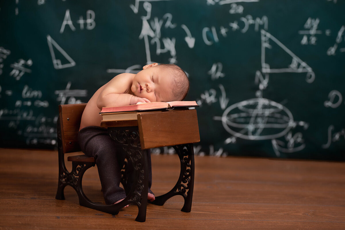 Adorable newborn posed in a classroom-themed setup with a tiny chalkboard, captured in Overland Park, Kansas. Perfect for families seeking creative newborn photography with unique themes in the Kansas City area.