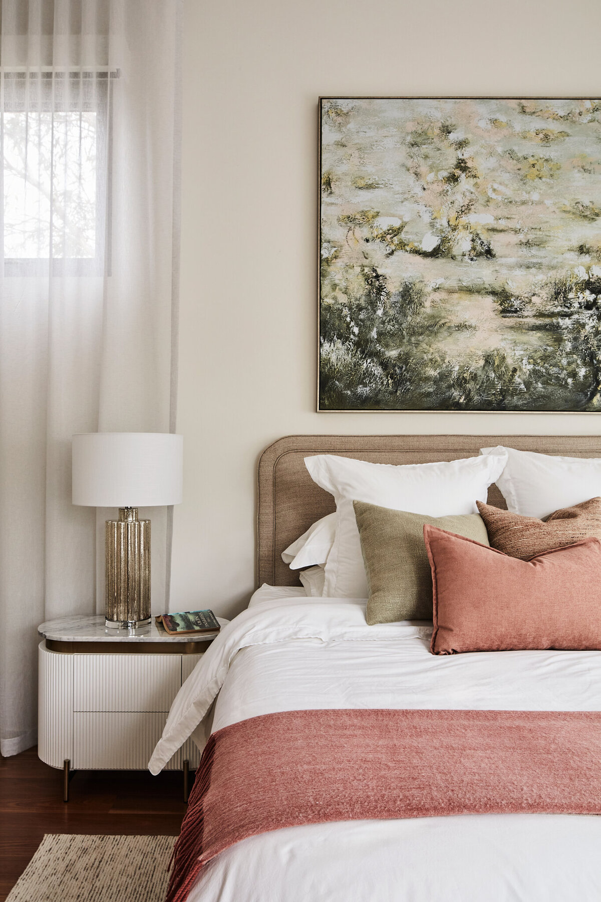 Modern residential bedroom interior in Melbourne with a neutral color palette, featuring a bed with a white comforter and blush-colored pillows. Gold-toned bedside lamp, contemporary wooden headboard, and a large abstract painting above the bed.
