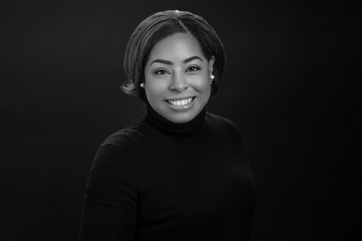 A woman with short natural hair, wearing a black turtleneck, smiling warmly against a dark background.