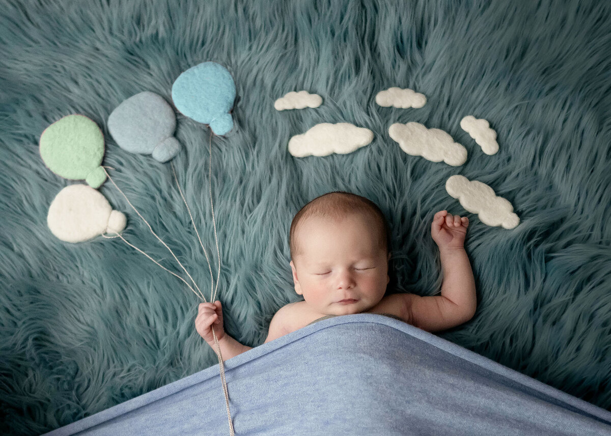 A newborn baby sleeps while holding balloon strings under clouds in the studio of a Pella Newborn Photographer