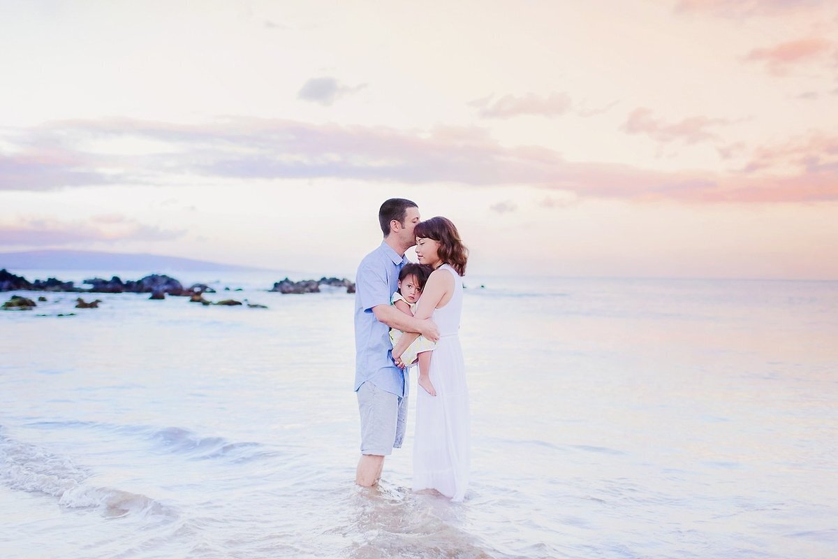 Sunrise portrait of husband kissing wife on the forehead with young child looking at the camera and Maui landscape in the background