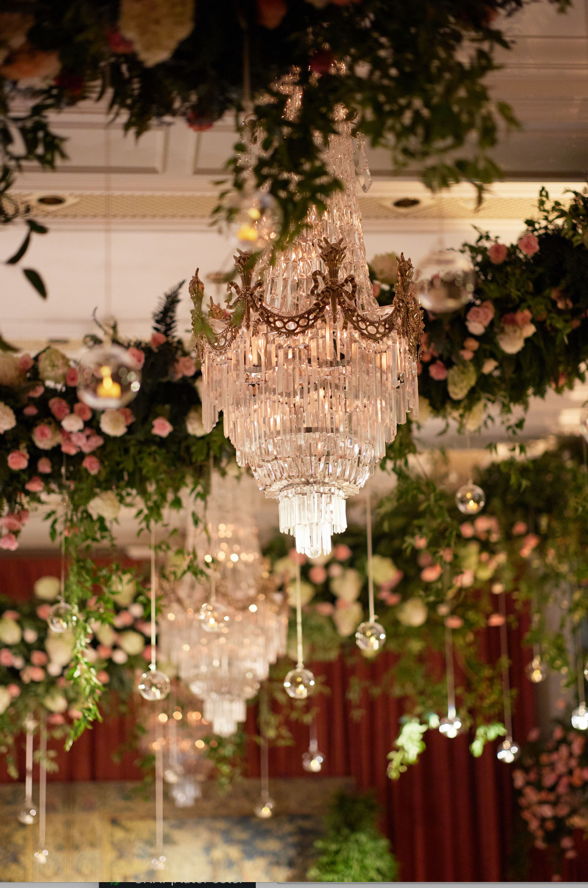 The photo was taken at the Willard Intercontinental in Washington D.C., which is renowned as one of the top wedding venues and hotels in the capital city. Captured by photographer Shawn Connell, with Clayton Films as videographers, the image showcases a luxurious chandelier adorned with crystal pendants, set amidst lush green foliage and a stunning array of pink and white flowers. Additionally, smaller hanging glass orbs with candles can be seen, adding to the elegant and enchanting ambiance of the scene.