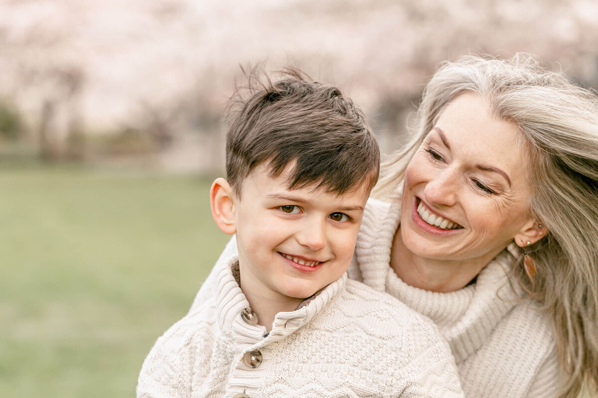 Mama  holding her 5 year old son on her lap and smiling at him with her hair blowing in the wind. Boy is smiling at the camera. They are sitting on a green lawn together with blooming cherry trees blurred out in the background. They are both wearing cream sweaters.