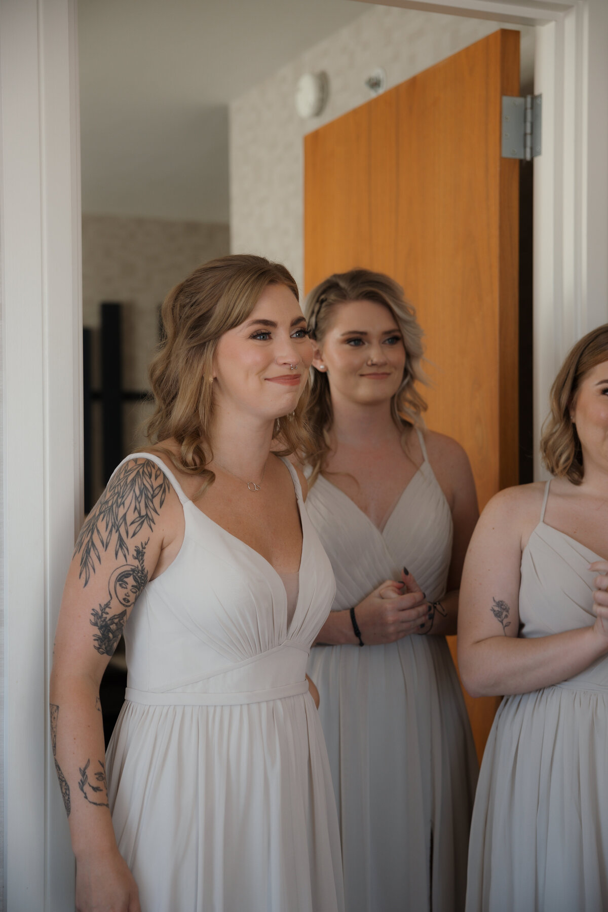 Bride sharing her first look with her bridesmaids, who are smiling and cheering as they see her wedding gown for the first time in an elegant room.