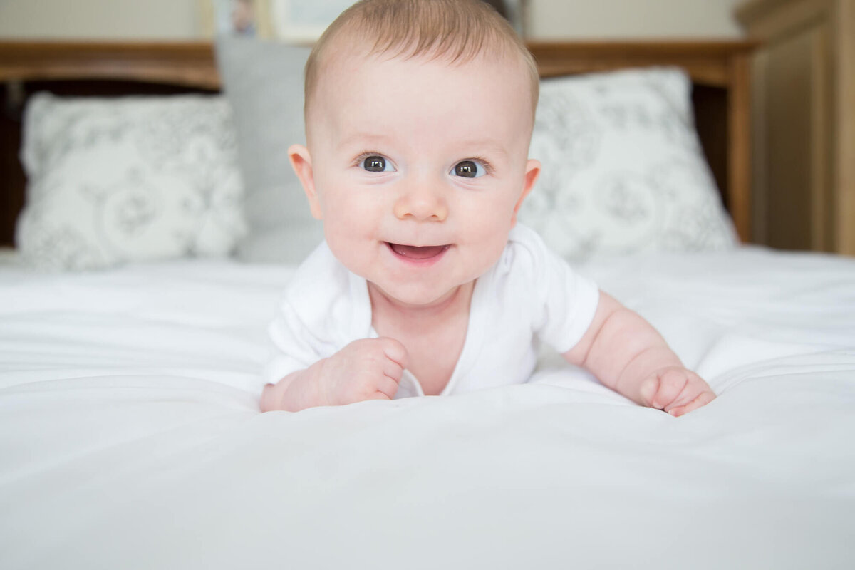 Excited infant boy in white shirt in a white bed holding his head up with a smile