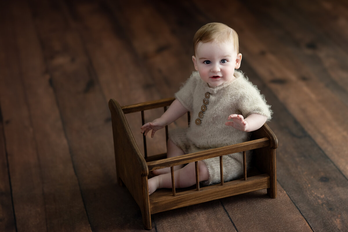  Rustic studio portrait of baby boy sitting in a wooden bed wearing a cozy knit romper—timeless textures and charm.