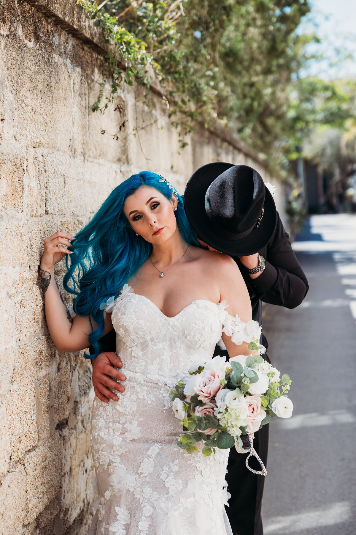 Bride with electric blue hair leans against a stone wall while groom kisses her shoulder in a sultry downtown St. Augustine portrait.