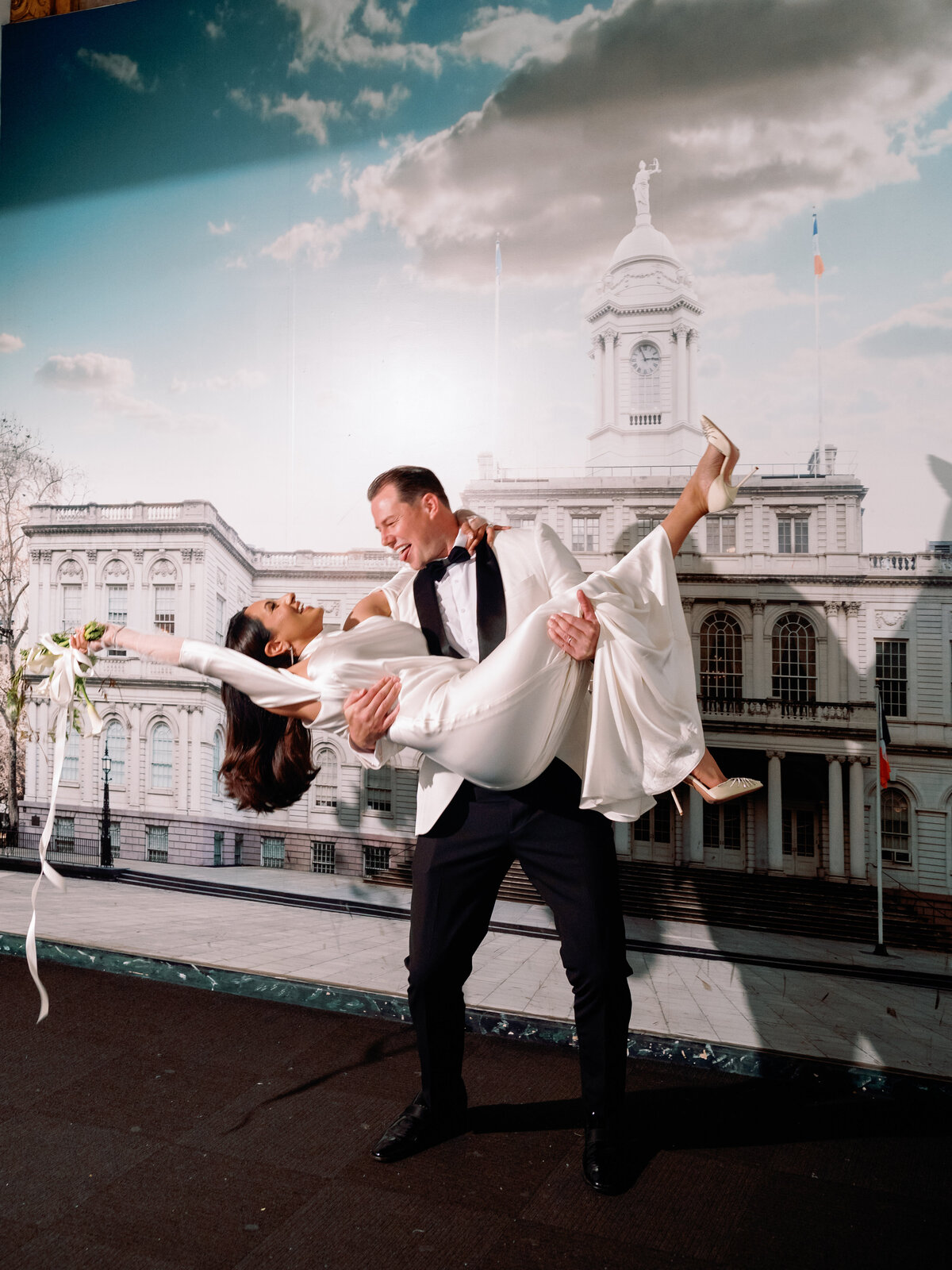 Groom Chris lifts bride Japna in front of New York City Hall, both smiling joyfully after their wedding ceremony.