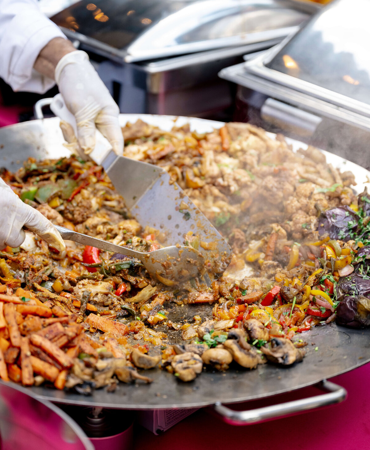 A chef wearing gloves cooks a variety of vegetables and meat on a large round grill. The dish includes mushrooms, peppers, and spices, and is being stirred with metal spatulas.