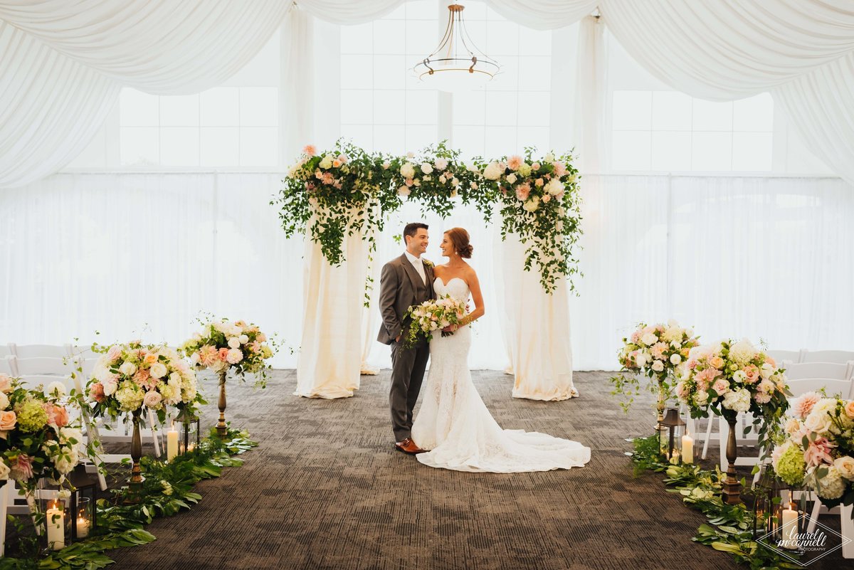 bride and groom at Newcastle Golf Club wedding standing in front of greenery chuppah, and aisle lined with tall flower arrangements in white tent