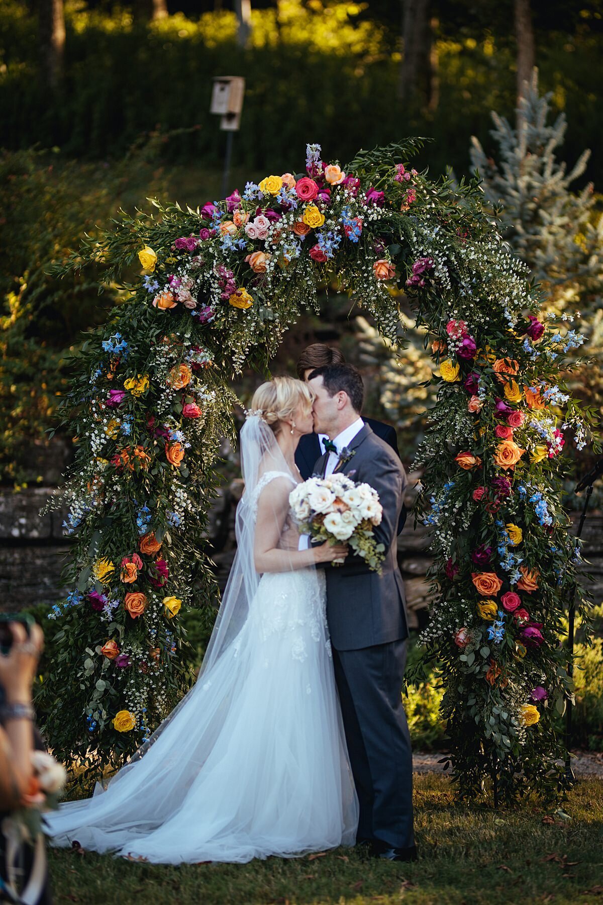 The bride, in a long sleeveless lace gown with a long sheer veil, holding a white, ivory, peach and blush bouquet kisses the groom in a charcoal gray tuxedo in front of a lush wedding arbor covered in greenery, red, yellow, orange, hot pink, blue, and purple flowers at Cheekwood Botanical Gardens