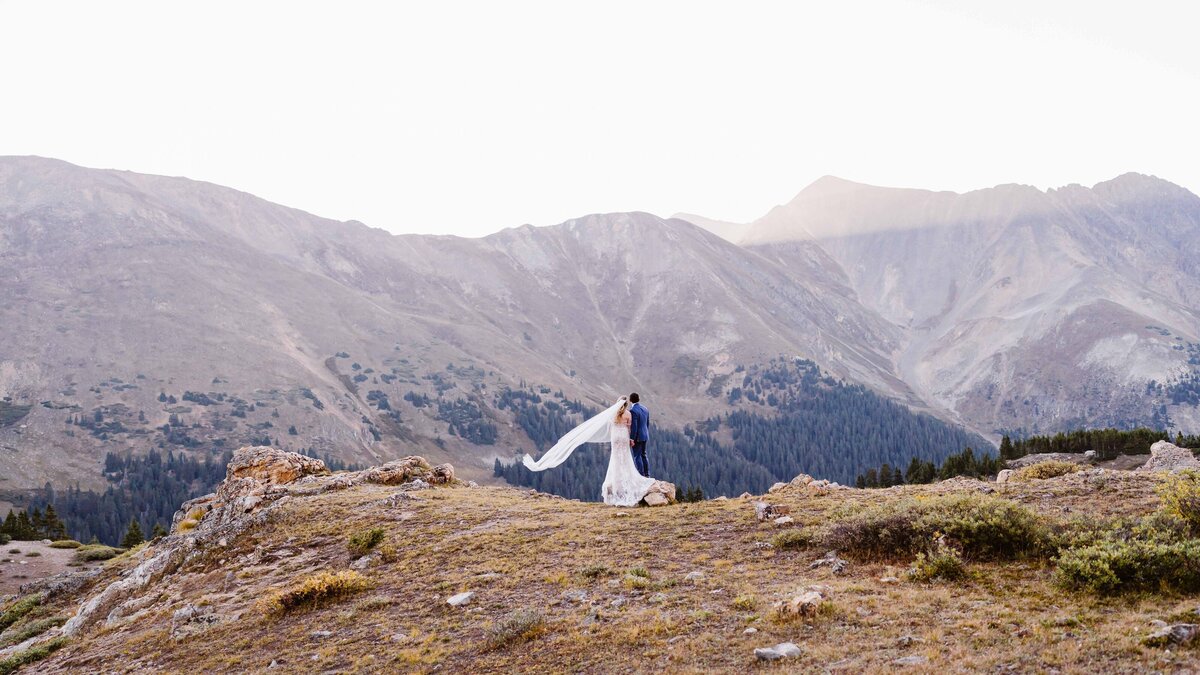 Bride and groom stand together at sunrise on a rocky ridge overlooking Pass Lake at Loveland Pass, Colorado, with the bride’s veil flowing in the mountain breeze.