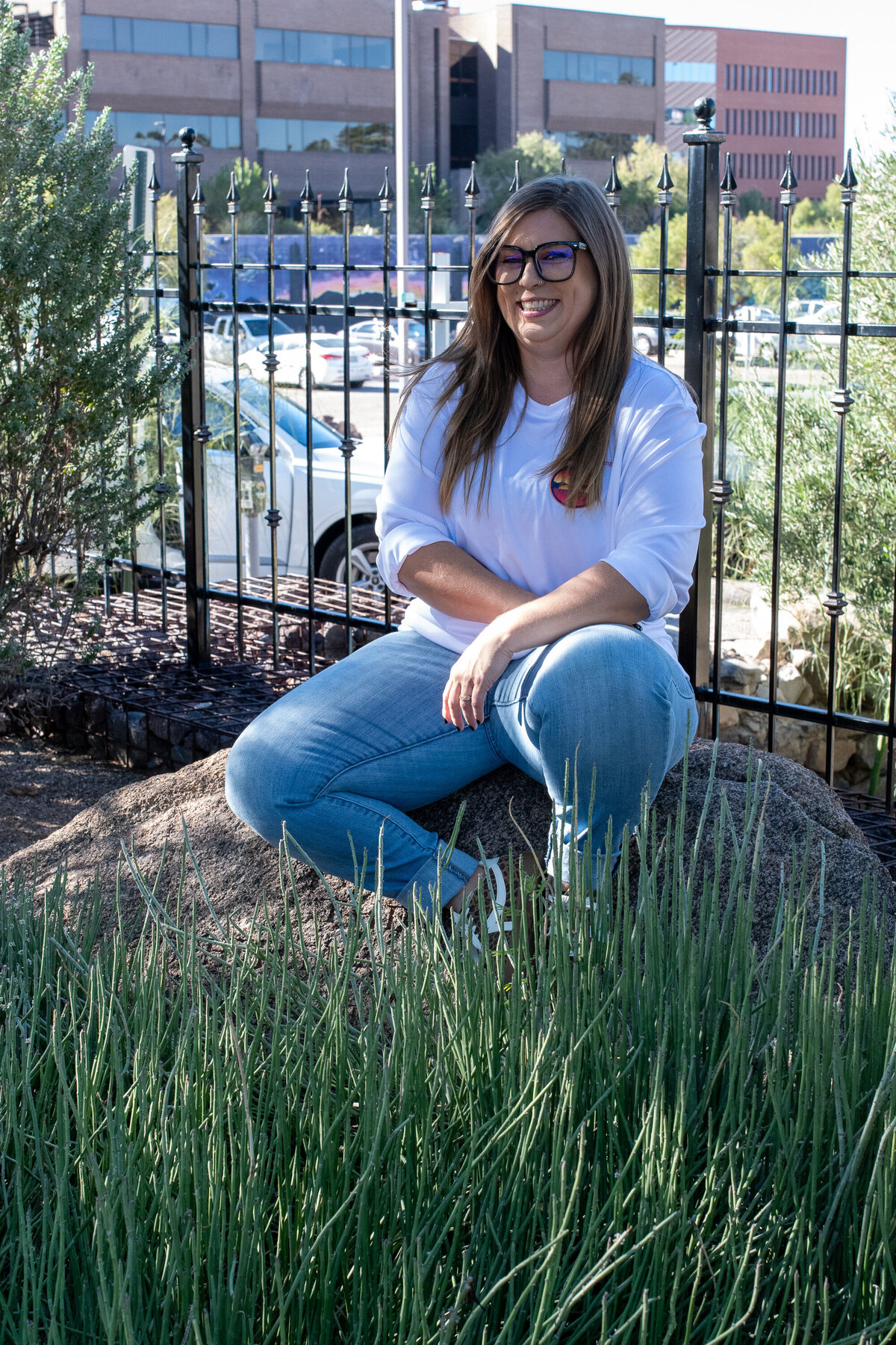 Woman sitting on a rock in a grassy outdoor area, smiling while wearing glasses and a white top, photographed by Vyrl Photo, showcasing Tucson brand photography.