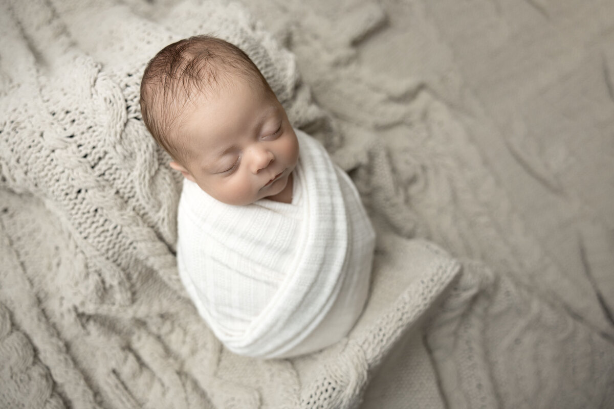 A swaddled baby sleeps soundly surrounded by a tan knitted blanket.
