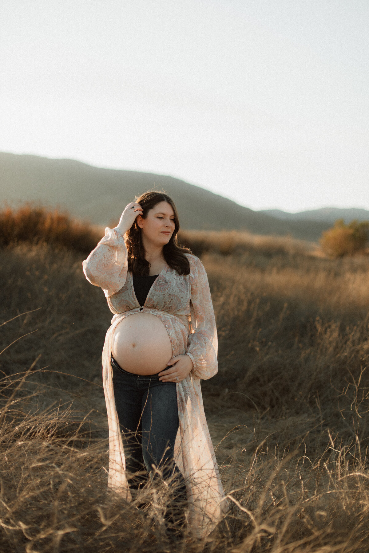 Pregnant woman standing in a sunlit field at golden hour, gently holding her bare belly and wearing a sheer floral robe over jeans, surrounded by soft grasses and distant hills.