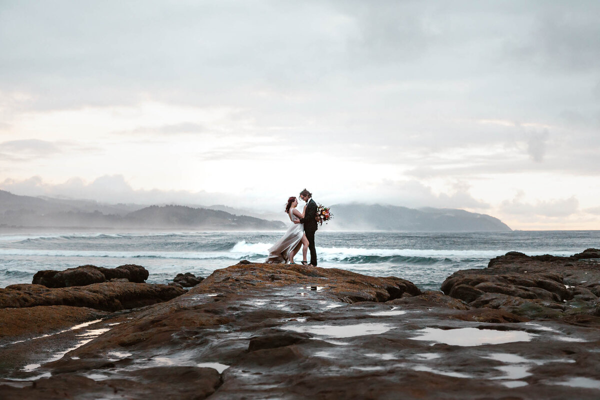 during their moody Oregon coast elopement, a bride and groom in their wedding attire embrace while standing atop a rocky shore. Wind makes the brides dress billow and the blue-gray waves roll behind them as a layer of fog rolls in.