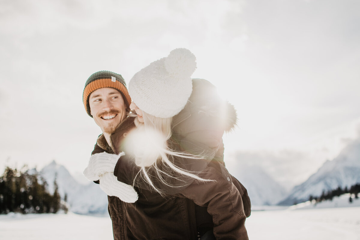 woman piggy bakcing on mans back with the sun beaming over her shoulder as she looks at his smiling face during their snowy winter engagement session by jackson hole photographers