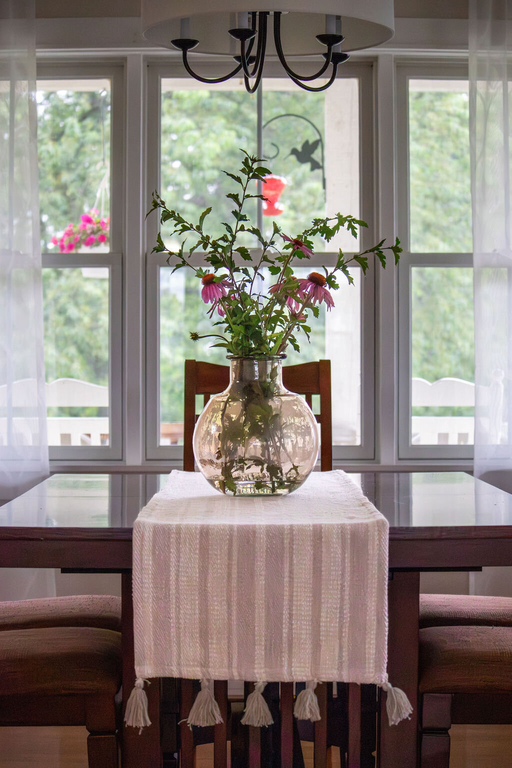 A dining table with a glass vase filled with purple coneflower stems and green leaves as a centerpiece, set on a white runner. The table is surrounded by chairs with striped cushions, and large windows with sheer white curtains reveal a view of lush greenery and a bird feeder hanging outside. A black chandelier hangs above the table.