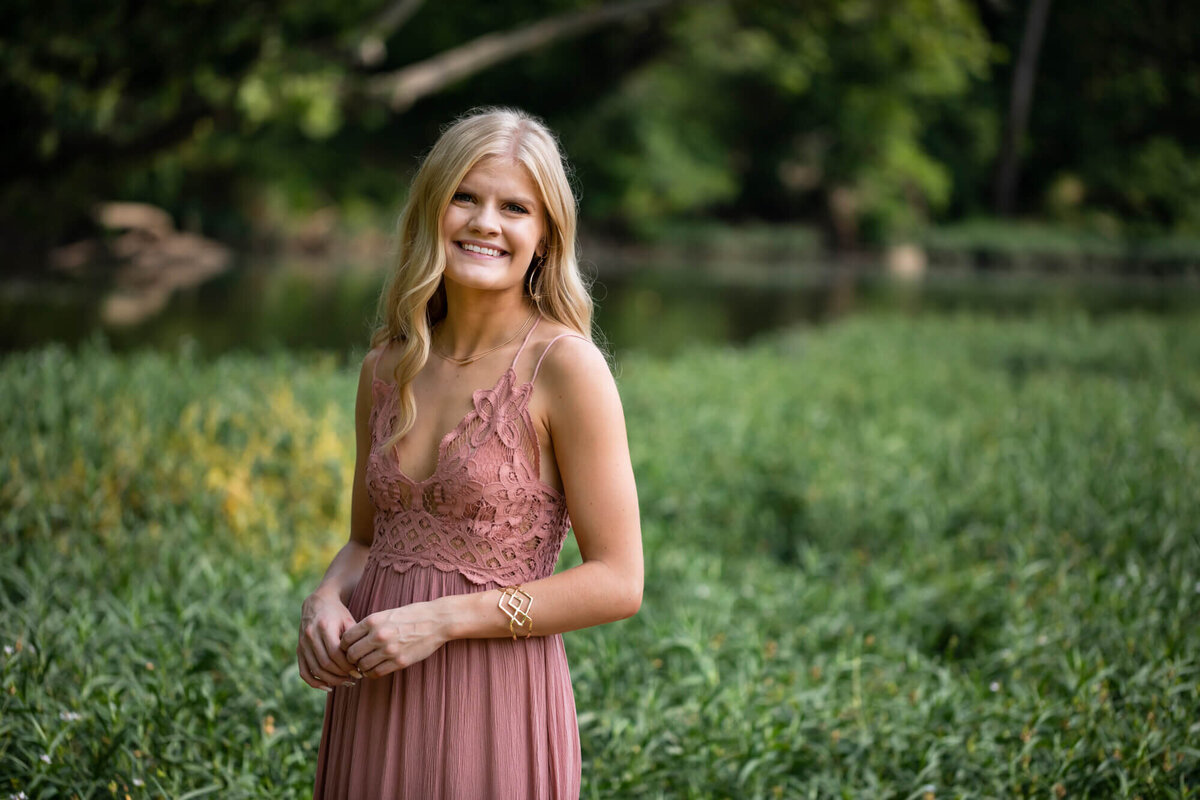 A beautiful blonde teen girl poses for a senior portrait wearing a dusty rose lace dress in front of a lush lake shore. Captured by Springfield, MO senior photographer Dynae Levingston.