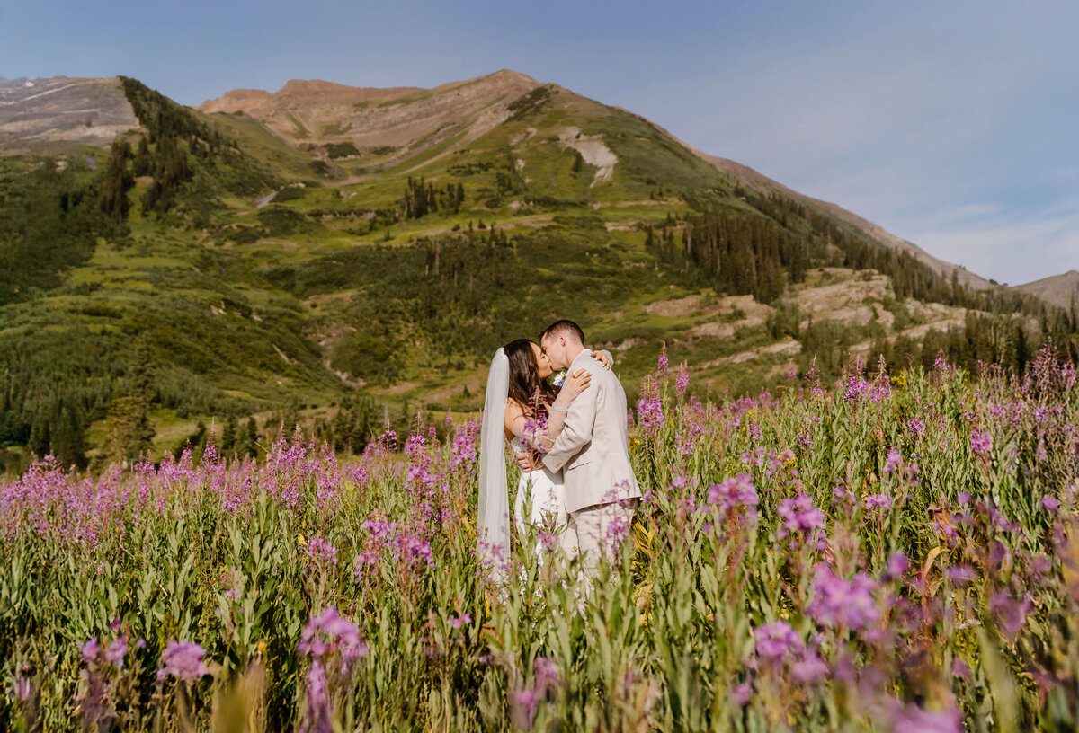 Bride and groom kiss in a field of purple wildflowers with mountain views during their summer elopement in Crested Butte, captured by Colorado elopement photographer Alpine Vows.