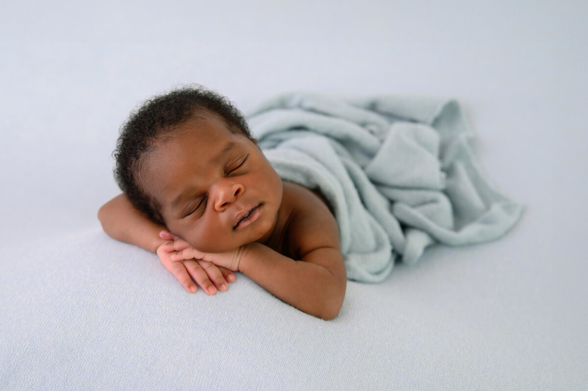 black newborn baby boy laying on a blue background with a blue wrap on him held tilted to the side on his hands for his Burlington, Ontario, newborn photography session.