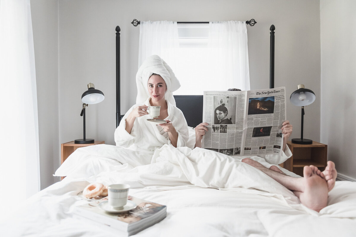 A woman wrapped in a towel robe sips coffee in bed while her partner reads the newspaper, surrounded by breakfast and soft morning light—a relaxed, editorial moment that feels quietly intimate and effortlessly chic.