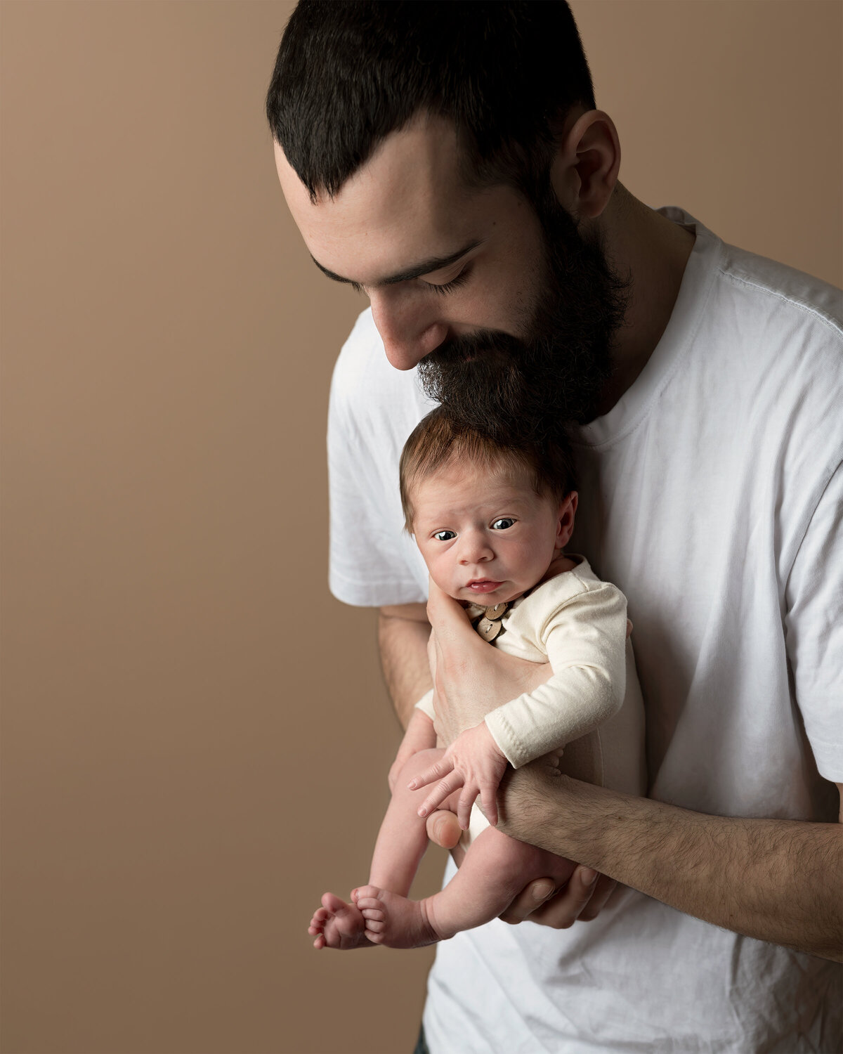 Emotive studio portrait of a bearded father tenderly holding his newborn baby in Paris, Ontario. Captured by Meraki Photographic Creations. Photographed in-studio at Meraki Photographic Creations in Paris, Ontario.
