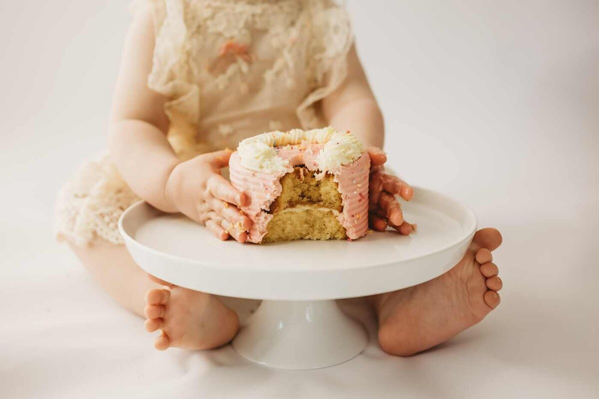 Ruua Hamdan Photography (UK): A baby in a lace dress sits with bare feet and holds a partially eaten pink and white cake on a white stand