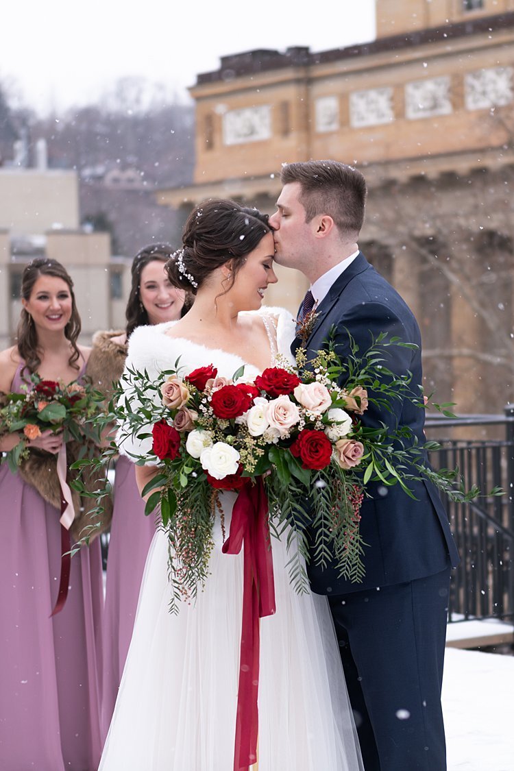 Groom kissing Bride's forehead on a snowy rooftop while Bridesmaids look on at University Club in Pittsburgh, PA