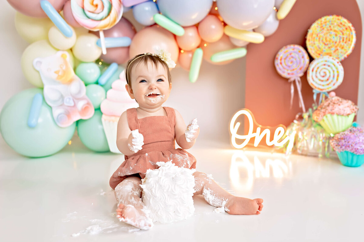 Toddler girl celebrating her first birthday. She is squeezing cake in her hands and smiling.