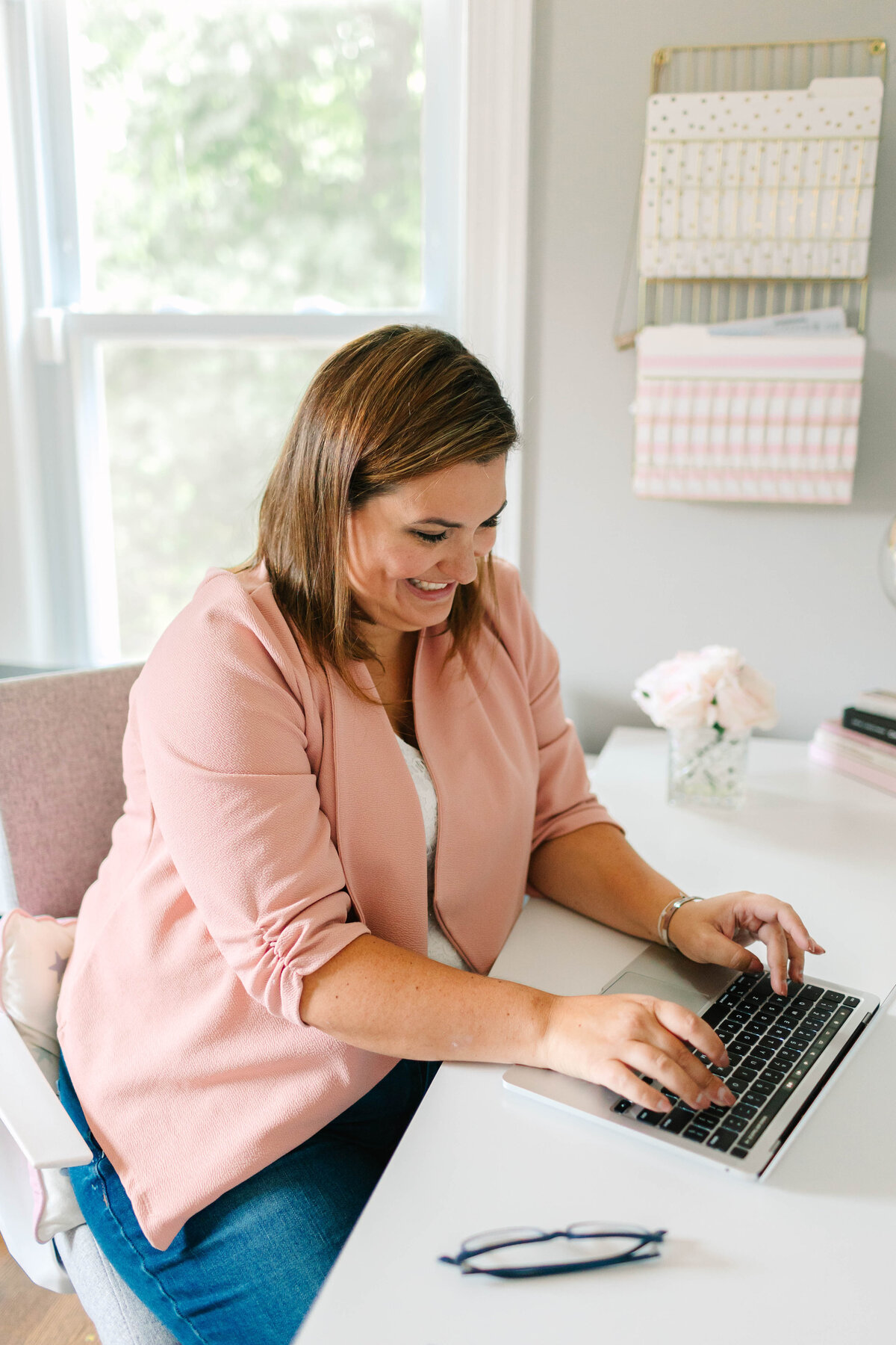 A smiling woman in a blush pink blazer types on a laptop at a bright, organized desk. Behind her, a neatly arranged wall organizer and soft pink decor create a cozy workspace.