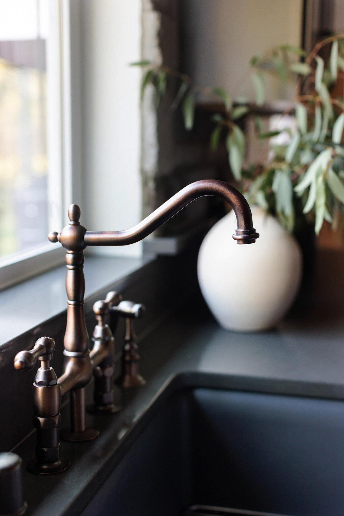 Dark grey kitchen sink with dark grey countertops, and antique bronze bridge faucet. Large beige vase with green florals next to the sink. Large window behind the sink. Antique bowl full of pears. Dish towel draped over the sink.