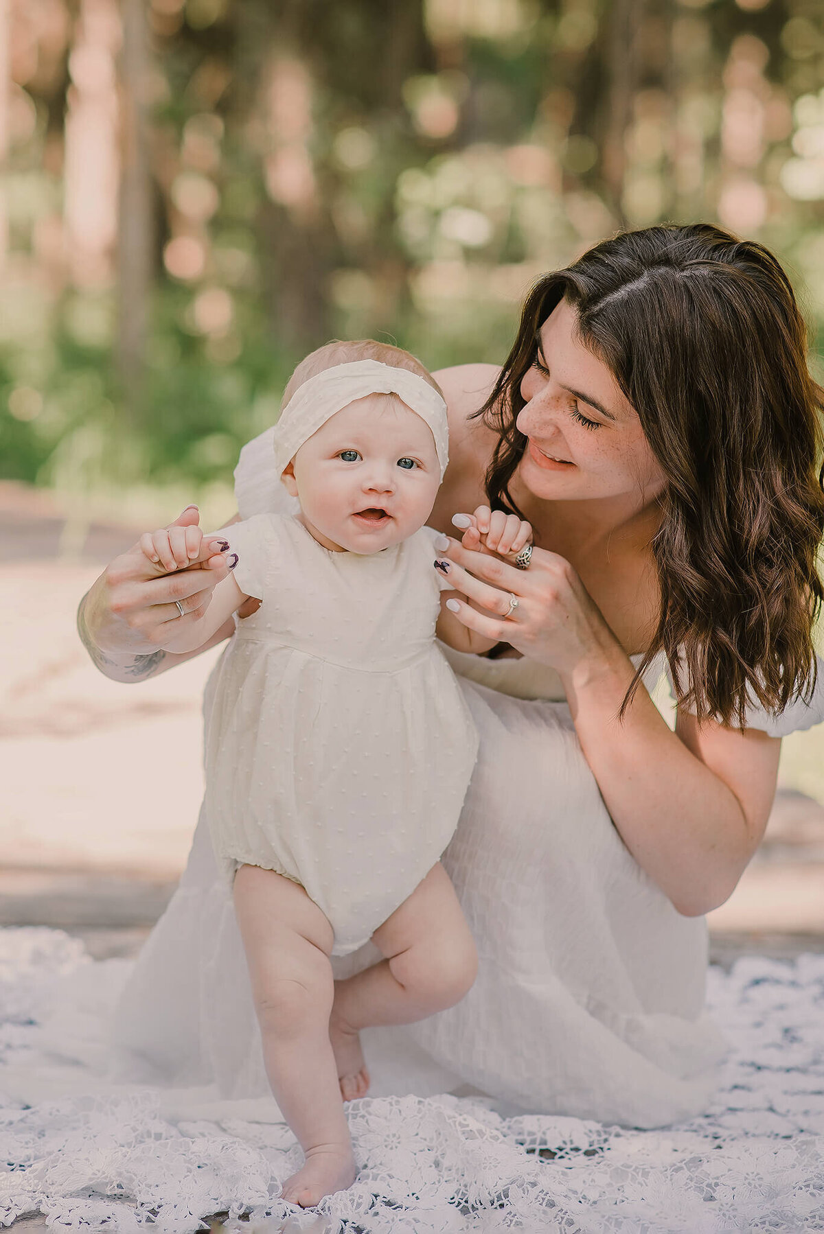 An up close portrait of a mother and daughter. They both wear white dresses and mom is crouched down holding baby girls hands while she stands. They both smile. Captured by Brittany Danielle