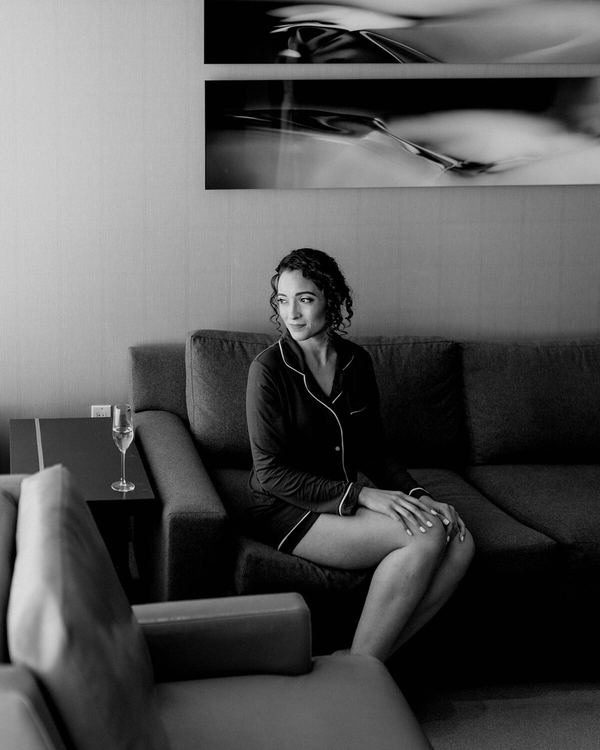 A contemplative black-and-white portrait of the bride sitting on a couch in dark pajamas, gazing off to the side, with a glass of champagne on a side table beside her.