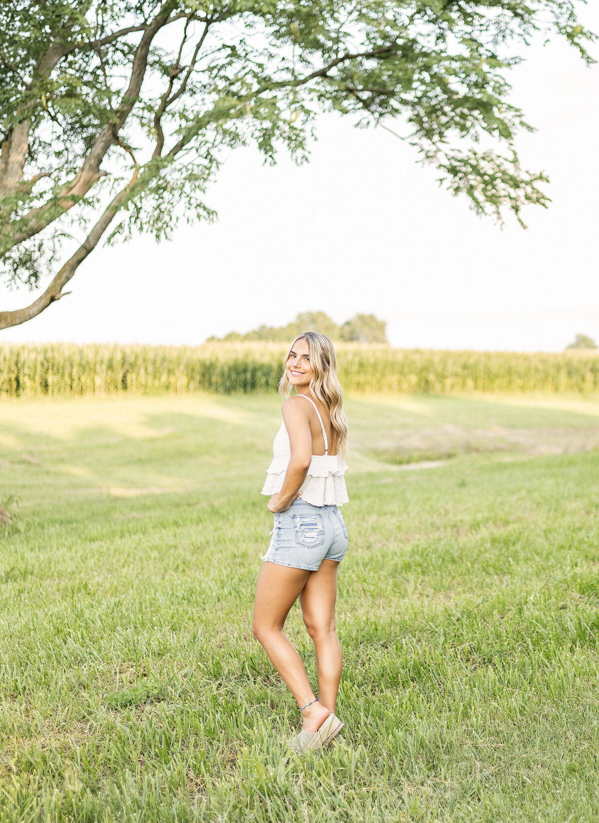 high school senior female, standing in grass with a tree and cornfield in the background, looking back at the camera smiling, wearing blue jean shorts and a white spaghetti strap top