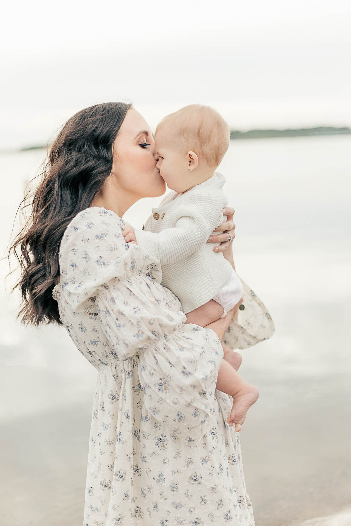 Mother in a floral dress holding and kissing her baby near the lake during a 6-month milestone session with her family in Kansas City.