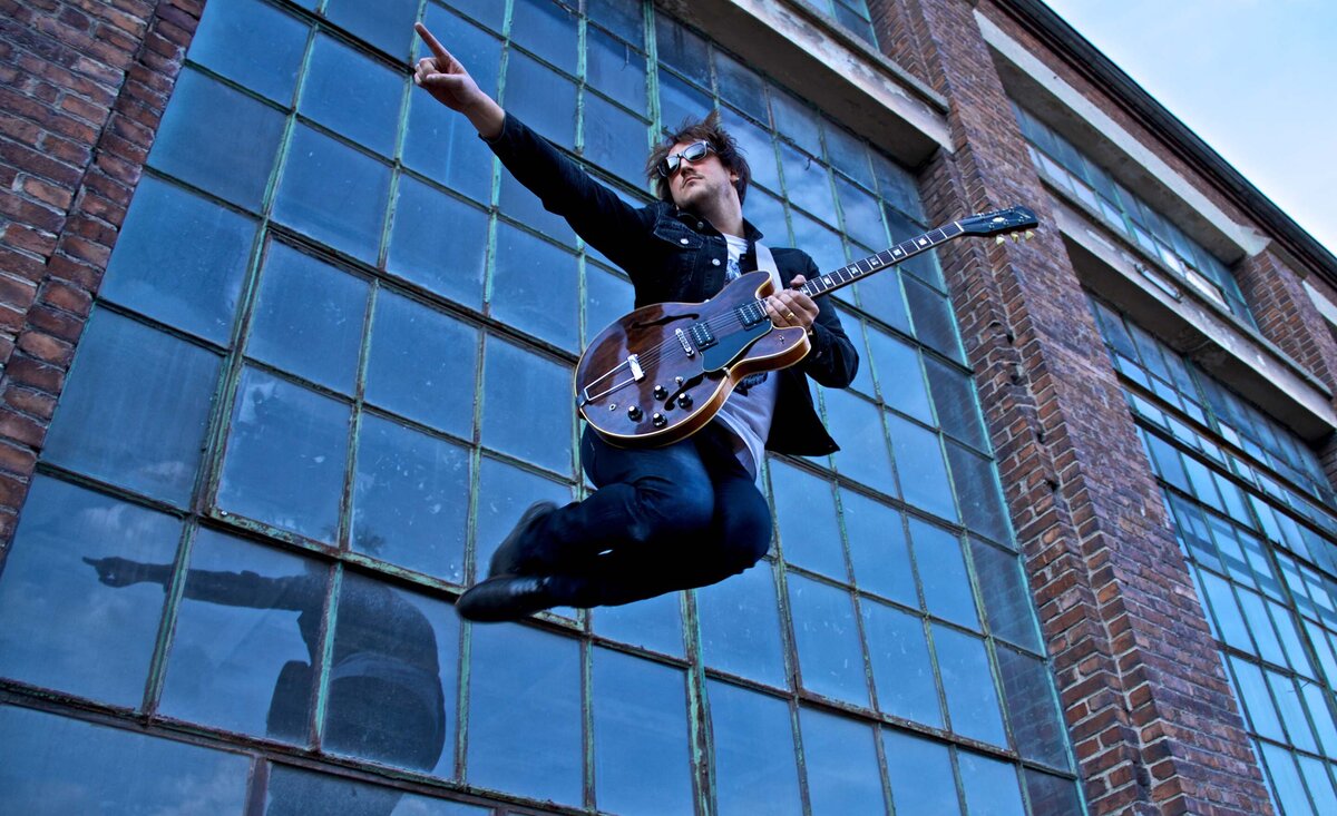 Male musician portrait Mikey Manville  wearing blue denim jeans jean jacket jumping while holding red electric guitar large window behind