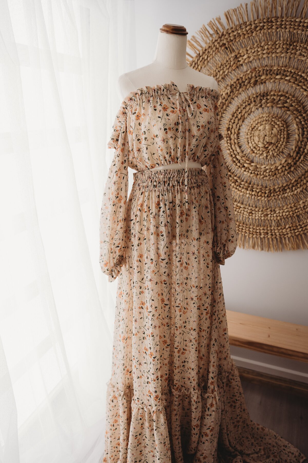 A floral off-shoulder dress is displayed on a dress form next to a window with sheer curtains, reminiscent of the elegant settings often captured by an NSW family photographer. A decorative woven wall hanging is visible in the background.