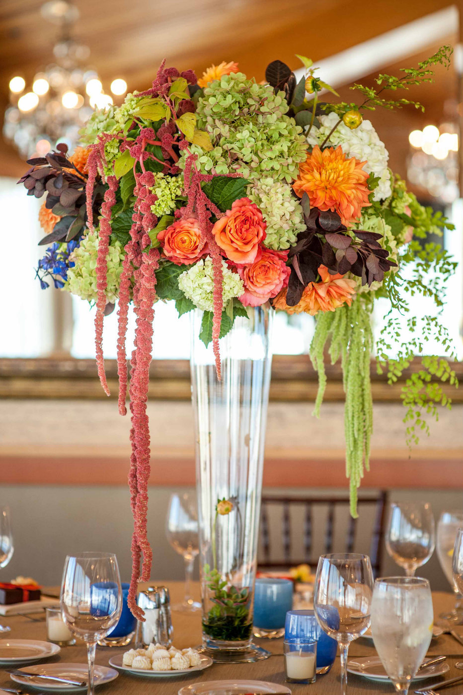 elevated centerpiece of  fall flowers: orange roses, green hydrangea, orange dahlias, ferns, hanging amaranthus on top of tall tapered glass vase with blue glassybaby candles
