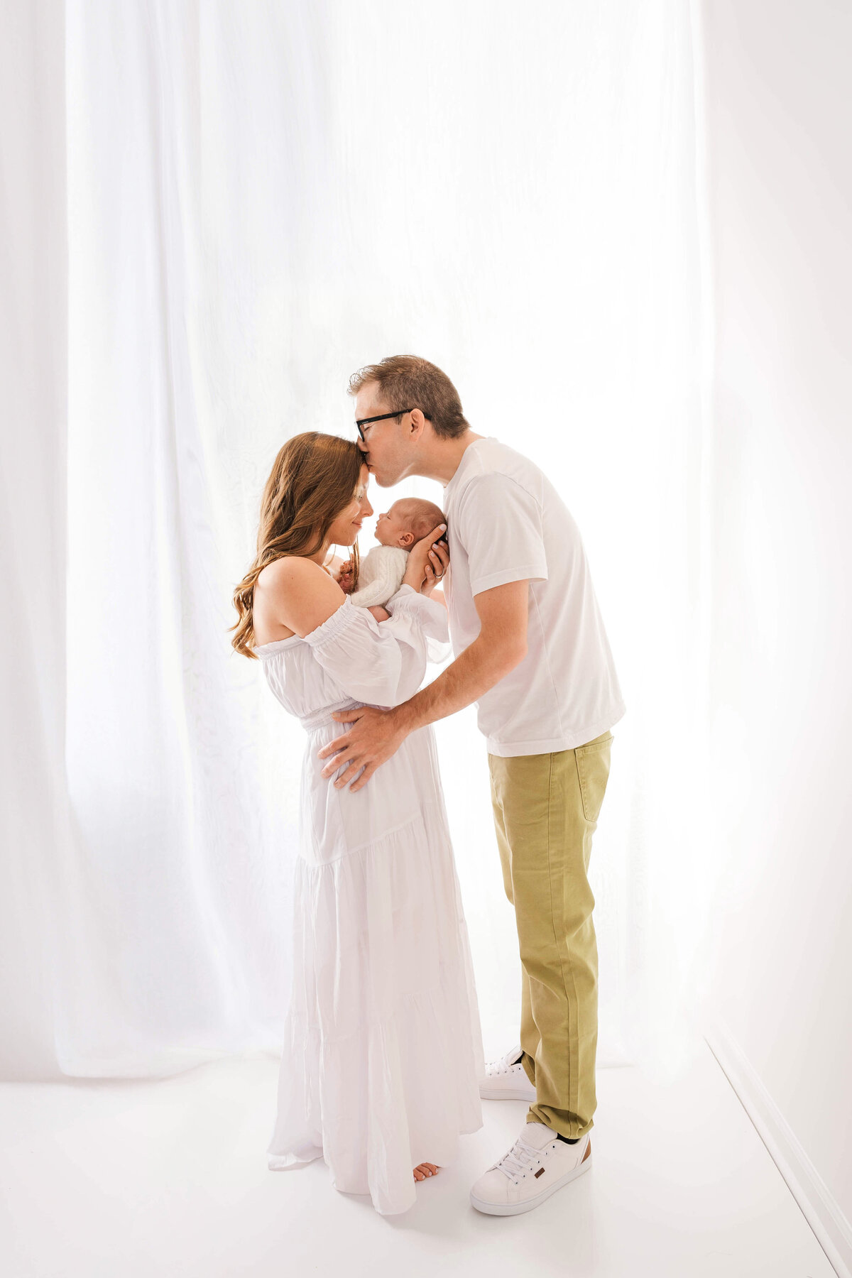 A husband kissing his sife's head as she cradles her newborn baby in front of a sheer curtain and window.