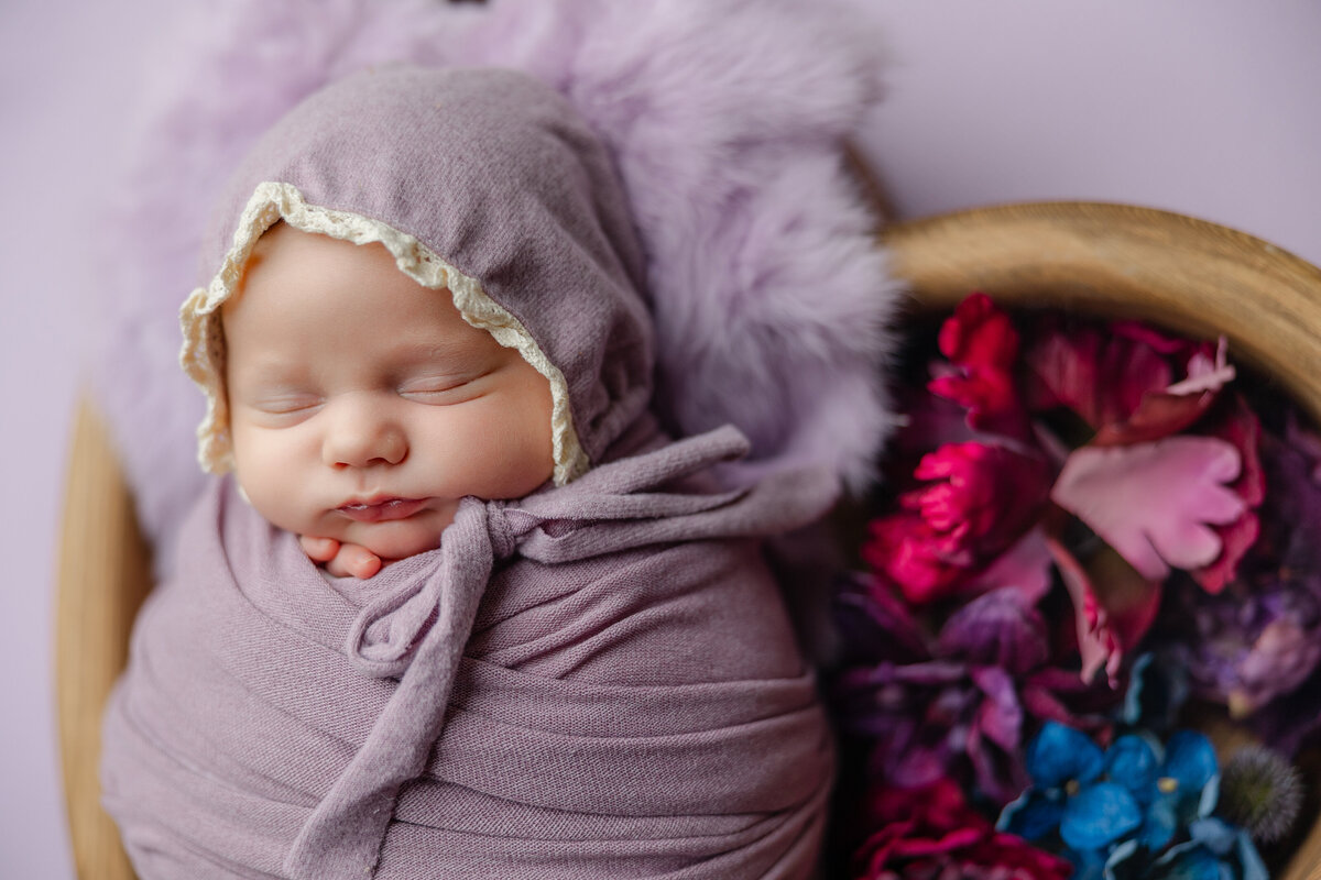 Close-up of a newborn baby girl wrapped in lavender with a lace-trimmed bonnet, resting beside bright florals and soft textures. Photographed by Inland Empire newborn photographer Kaitlyn Dawn