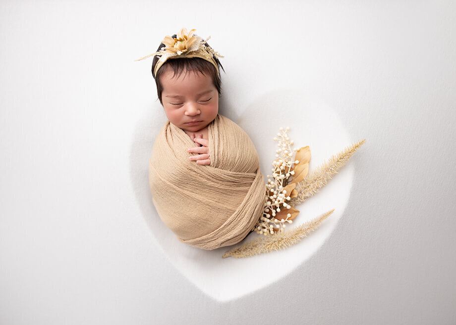 Female baby placed in a heart bowl underneath white fabric.  Baby is swaddled in  a toasted walnut color with nude and white florals with a matching headband