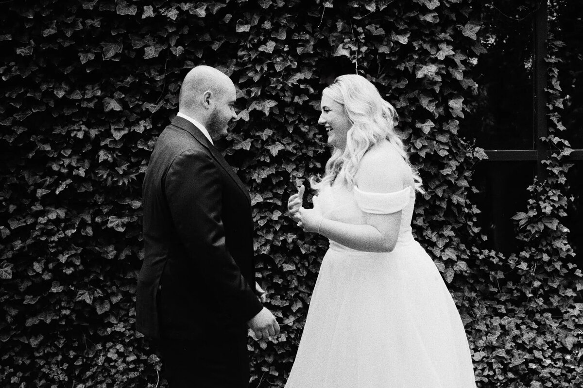 A bride and groom smile joyfully at each other in front of a leafy wall. Captured candidly by an NJ wedding photographer, the bride stuns in an off-the-shoulder white dress while the groom looks dapper in his suit.
