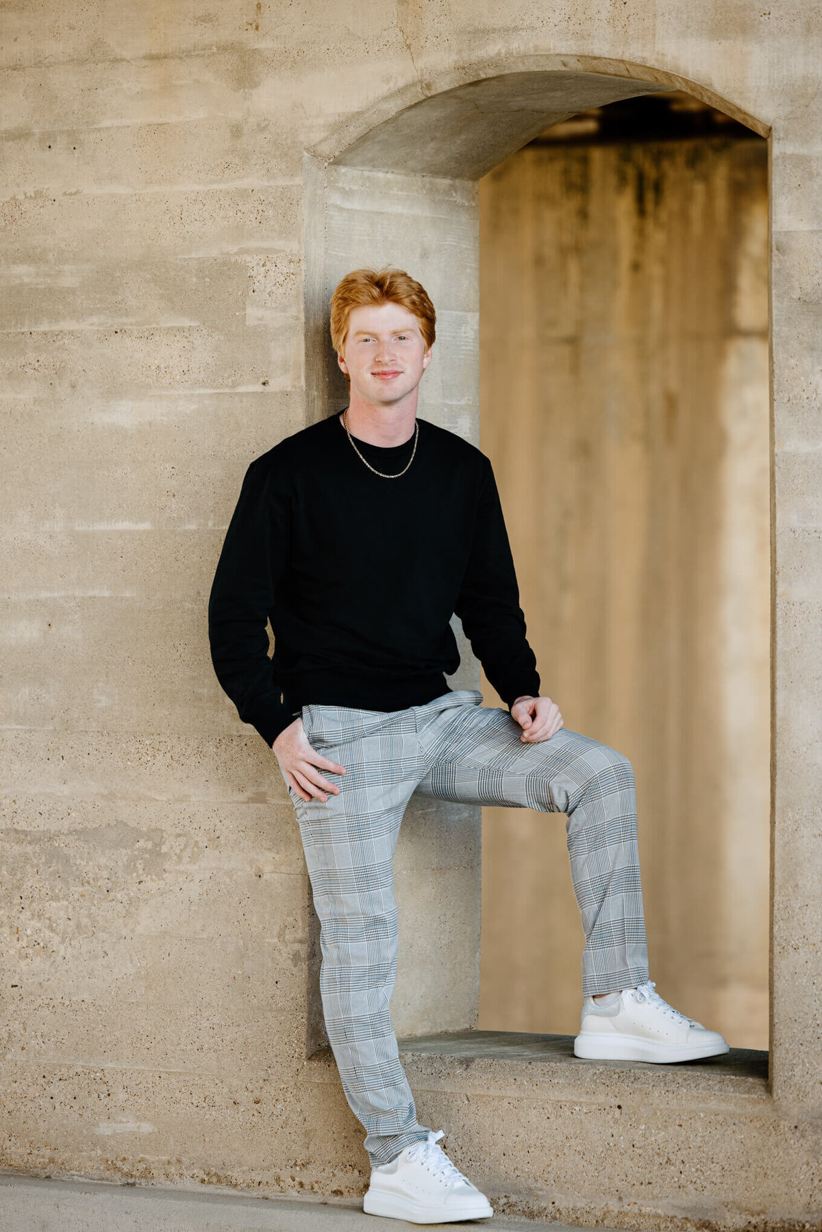 Senior portrait of graduate in black long sleeve shirt, plaid pants, and white shoes leaning on concrete wall in downtown Longview, TX