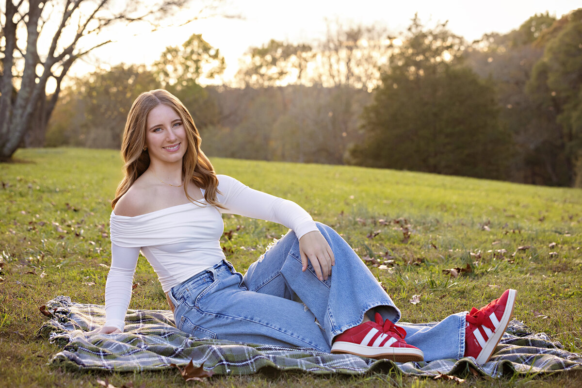 High school senior in jeans and white top sitting on grassy hillside at Anne Springs Close Greenway